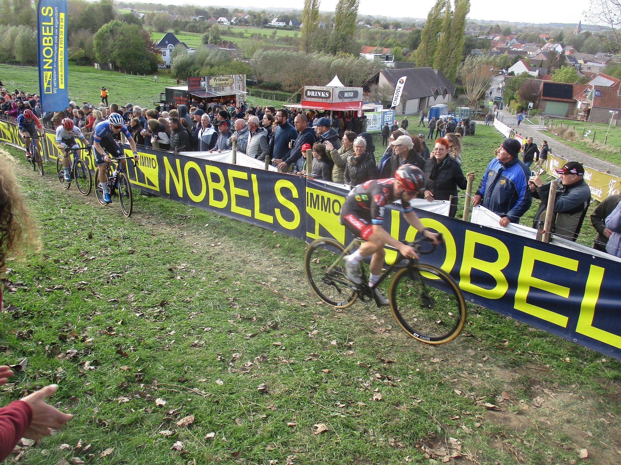 Eli Iserbyt mène la course sur une pente herbeuse le long de banderoles jaunes, suivi par d'autres coureurs, le public derrière les barrières.