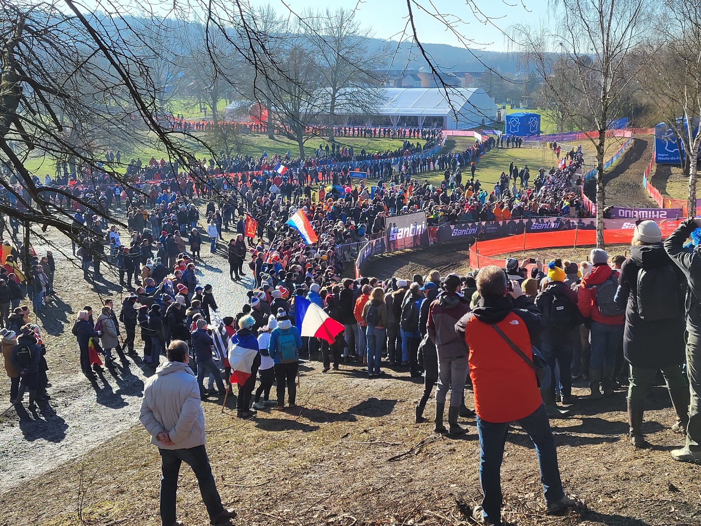 Vue en surplomb sur une foule de spectateurs et de drapeaux français massés le long du parcours, dans le parc boisé de Liévin.
