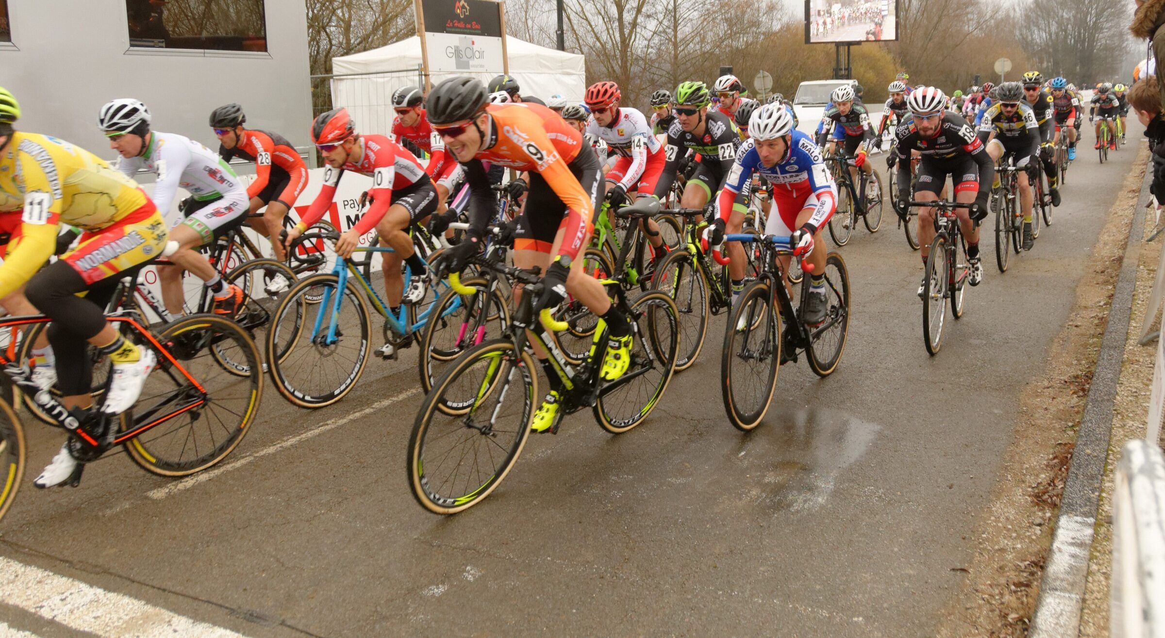 Le peloton serré des élites hommes roule sur une route mouillée, coureur en maillot jaune en tête de la file de gauche.