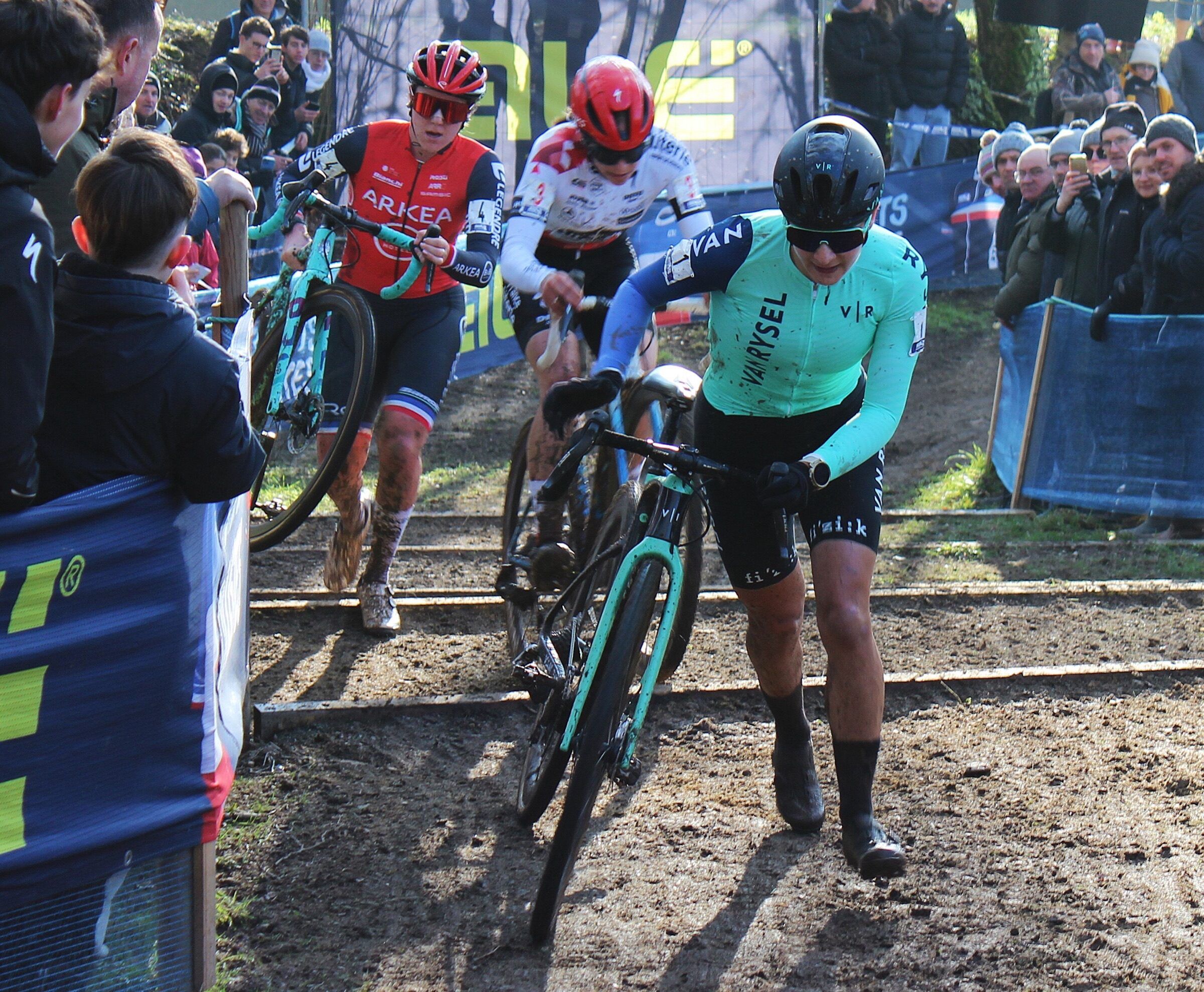 Trois coureuses franchissent à pied des marches de terre et de bois, vélo poussé ou porté, entre deux rangées de spectateurs serrés derrière les barrières.