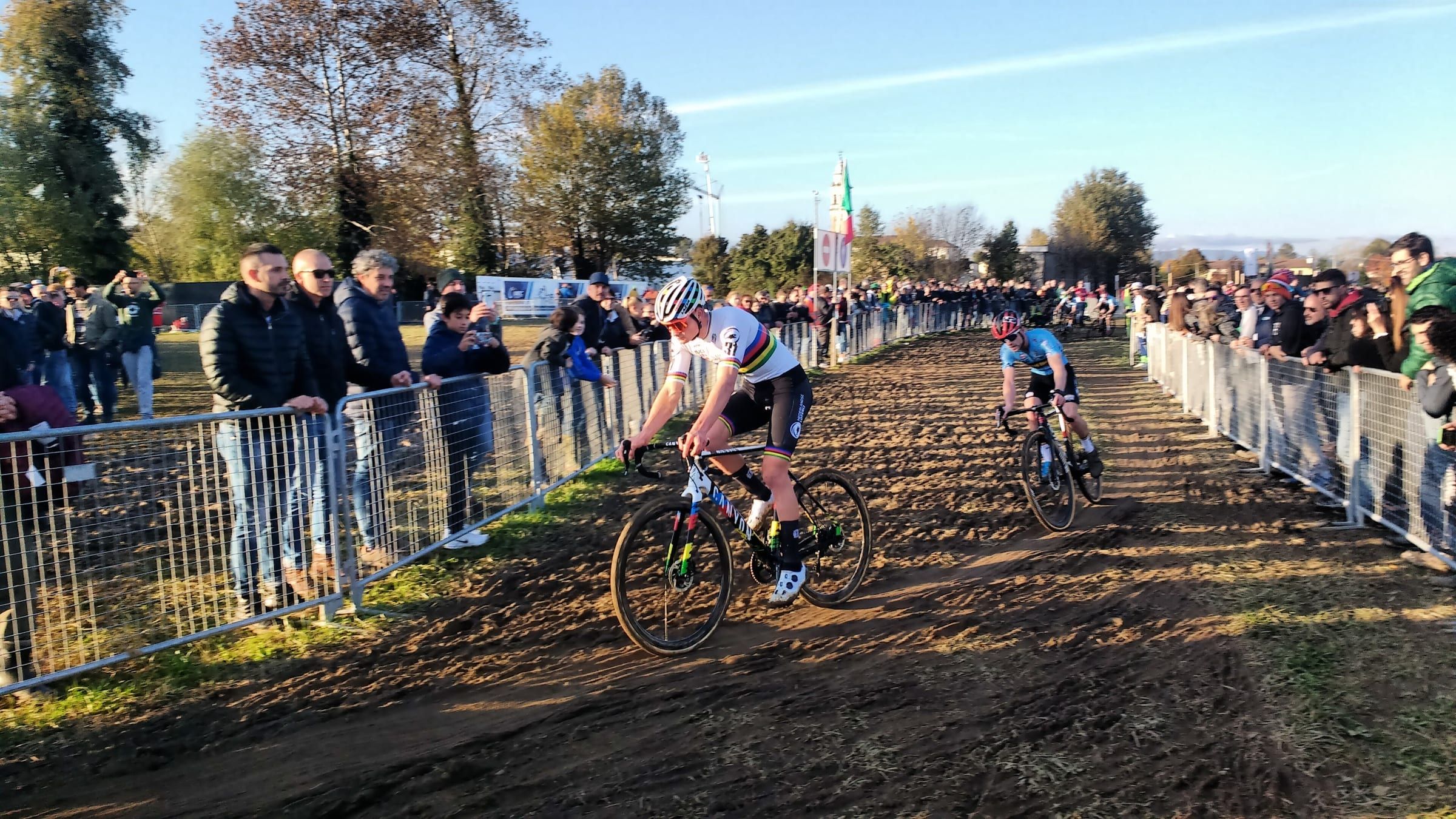 Un coureur en maillot arc-en-ciel pilote dans une tranchée de terre entre deux rangées de spectateurs, en plein soleil.